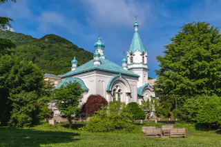 A white church with teal domes and spires sits among lush green trees, with benches on the grass in the foreground and forested hills and blue sky in the background.