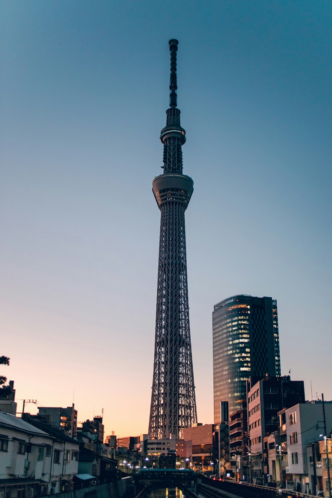 A tall broadcasting and observation tower stands prominently against the evening sky, surrounded by smaller buildings. The sky exhibits gradients of blue and peach, suggesting a sunset or sunrise. The tower features a lattice design near its base and a central observation deck.