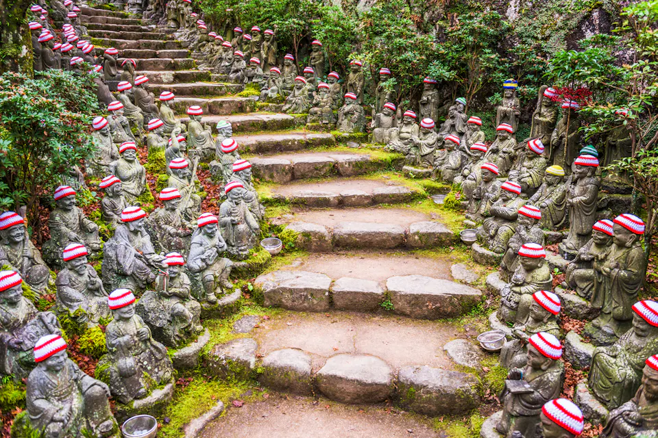 A stone path lined with small Buddhist statues, each wearing a red and white knitted hat. The statues are set amidst lush green foliage, creating a serene and peaceful atmosphere. The path ascends through stone steps surrounded by the statues on both sides.