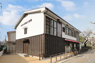 A two-story building with white walls and dark wooden accents stands on a quiet street, featuring large windows, a red awning, and Japanese writing on the facade beneath a blue sky with scattered clouds.
