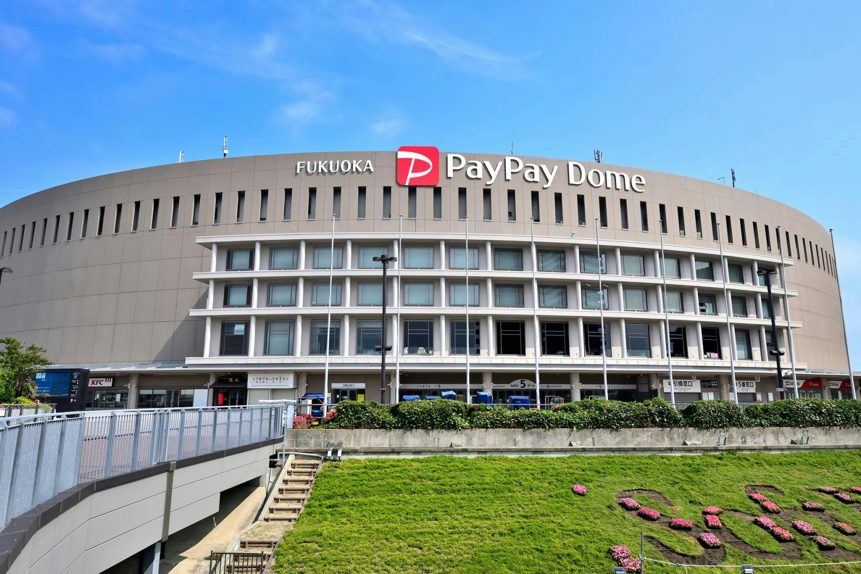 The exterior of Fukuoka PayPay Dome, a large, round stadium with the venue’s name and logo on top, under a blue sky. In front, there are stairs, a grassy area, and some flower arrangements.