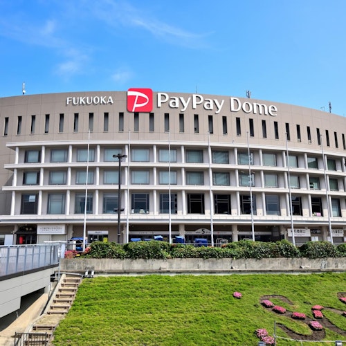 Fukuoka PayPay Dome The exterior of Fukuoka PayPay Dome, a large, round stadium with the venue’s name and logo on top, under a blue sky. In front, there are stairs, a grassy area, and some flower arrangements.