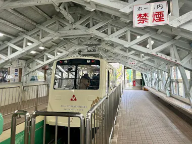 A small cream-colored cable car labeled "Ama no Hashidate Kasamatsu Park" is parked at a covered station platform with metal beams and Japanese no smoking signs overhead. Passengers are visible inside the car.