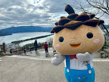 A large, smiling mascot in a pinecone costume poses on a scenic overlook with mountains, a river, and a cloudy sky in the background while tourists take photos.