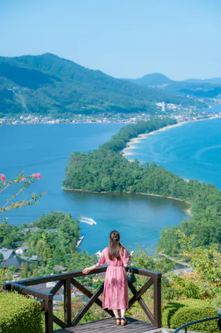 A woman in a pink dress stands on a wooden lookout, gazing at a scenic, tree-covered sandbar stretching across blue water with mountains and a village in the background under a clear sky.