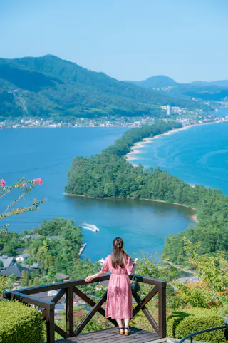 A woman in a pink dress stands on a wooden lookout, gazing at a scenic, tree-covered sandbar stretching across blue water with mountains and a village in the background under a clear sky.