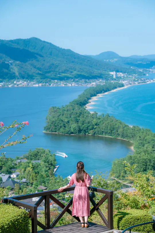 A woman in a pink dress stands on a wooden lookout, gazing at a scenic, tree-covered sandbar stretching across blue water with mountains and a village in the background under a clear sky.