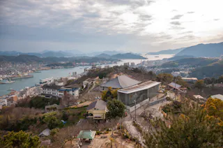 A scenic view overlooking a coastal town with a harbor, buildings, and roads nestled on a hillside. Surrounding mountains and the hazy sky provide a picturesque backdrop. Several large buildings and traditional structures are visible in the foreground.