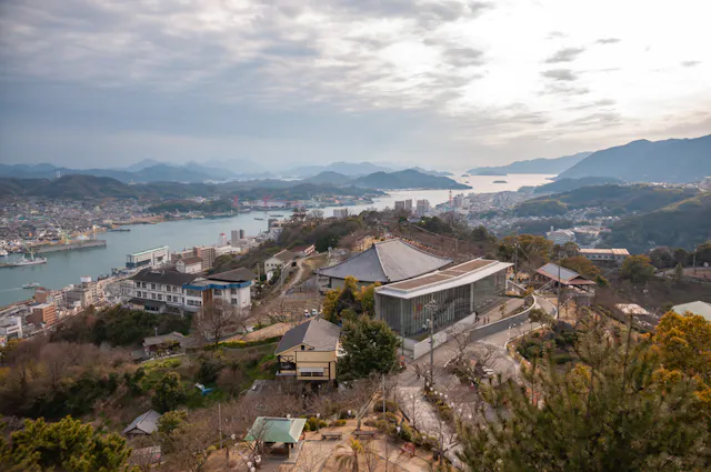 A scenic view overlooking a coastal town with a harbor, buildings, and roads nestled on a hillside. Surrounding mountains and the hazy sky provide a picturesque backdrop. Several large buildings and traditional structures are visible in the foreground.