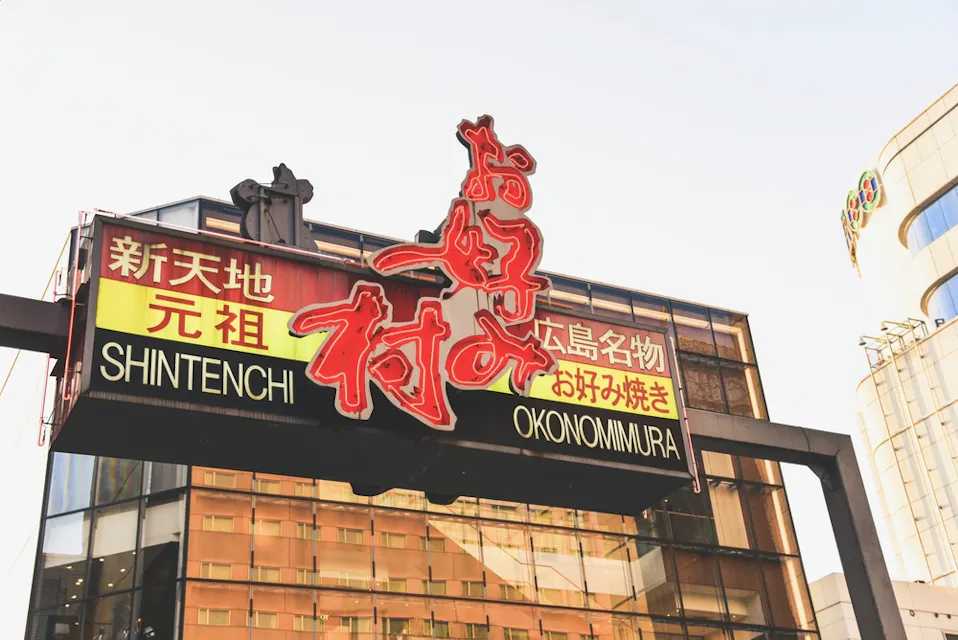 Okonomimura 在一座城市中,一个大型、色彩斑斓的标志上写着“新天地御好烧村”,并有日文字符。这个标志宣传一个以御好烧而闻名的地方,背景中可以看到高耸的建筑和晴朗的天空。