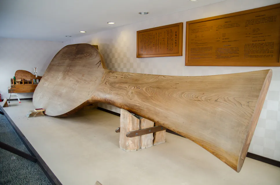 Largest Rice Scoop on Miyajima Island 这幅图描绘了一个大型木制清酒杯,展示在博物馆环境中。杯子是倒置的,底部支架抬高。杯子后面的墙上可见带有文字的展牌和各种小型展品。