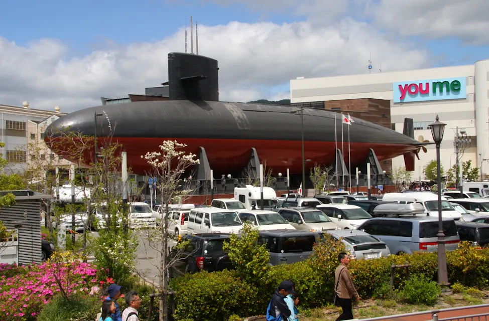 Iron Whale Museum 一艘大型黑红色潜艇展示在城市地区的陆地上,附近有人行走。背景显示了一栋"你我"大楼和一个停满汽车的停车场。前景中可见树木和开花植物,天空部分多云。