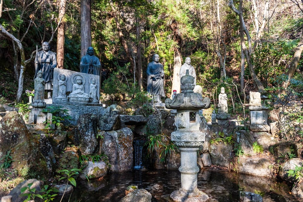 A serene outdoor scene featuring several stone Buddha statues arranged amidst lush greenery and trees. A small water feature with a gentle stream cascades over rocks in the foreground, and a traditional stone lantern stands prominently in the center.