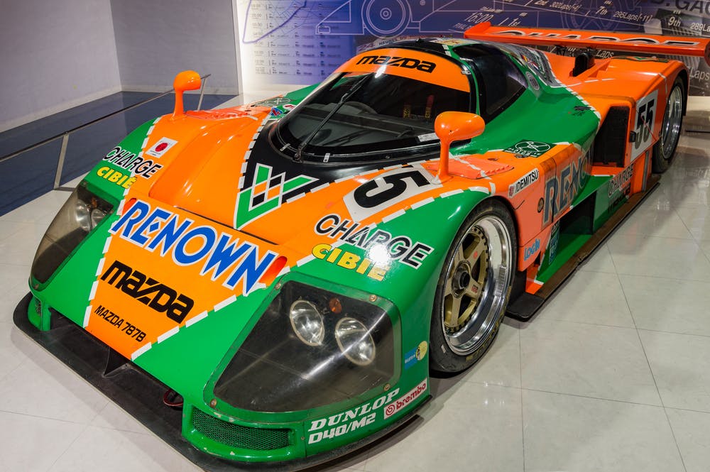 A vibrant green and orange Mazda 787B race car is on display. The car features various sponsor logos, including Renown and Charge, and has the number 55 on the hood and sides. The car has a sleek aerodynamic design and is displayed indoors on a polished floor.