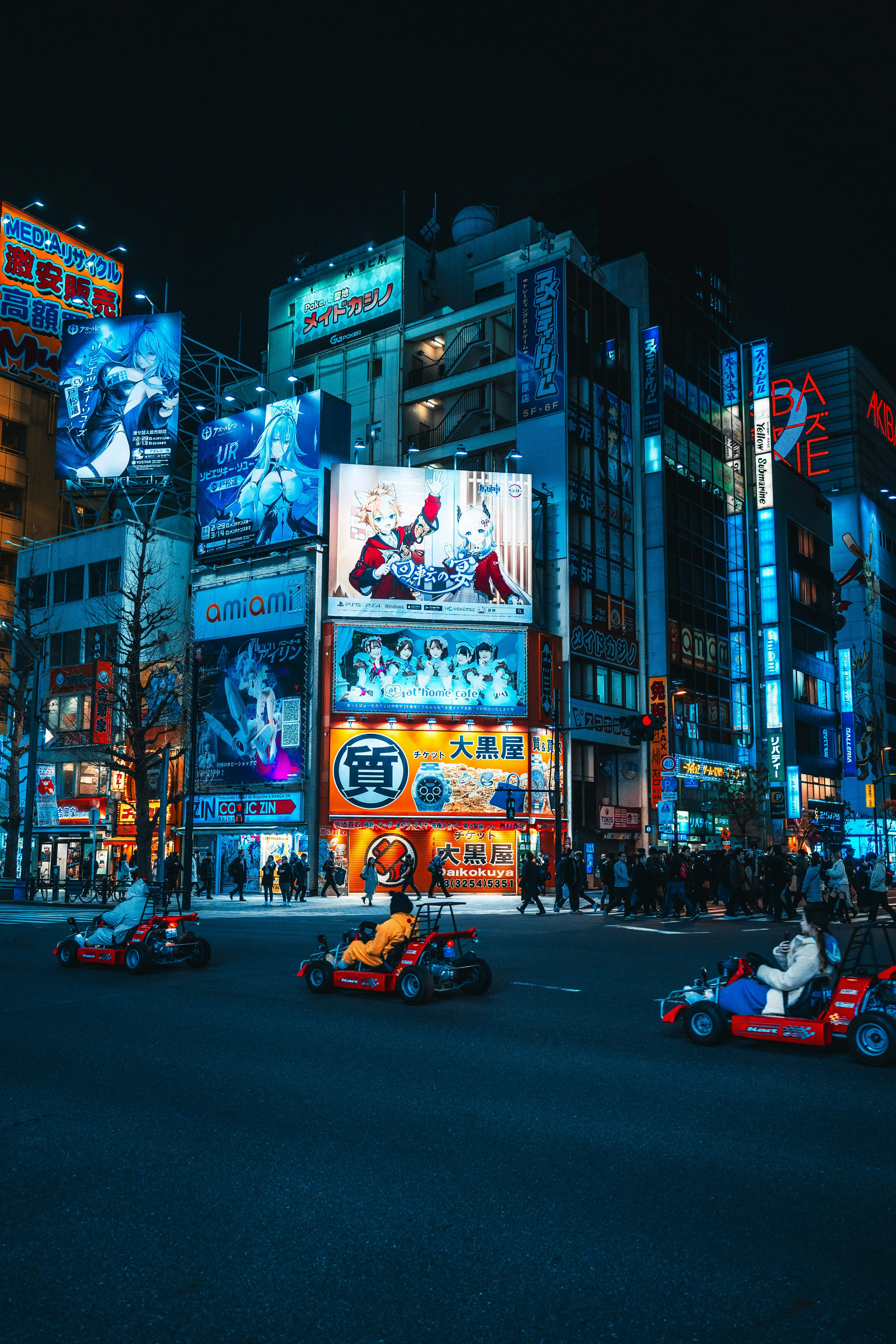 Nighttime scene of a bustling city street featuring several small go-karts driving in the foreground. The background is illuminated with colorful neon signs and advertisements on tall buildings. Many people are walking on the sidewalks, adding to the lively atmosphere.