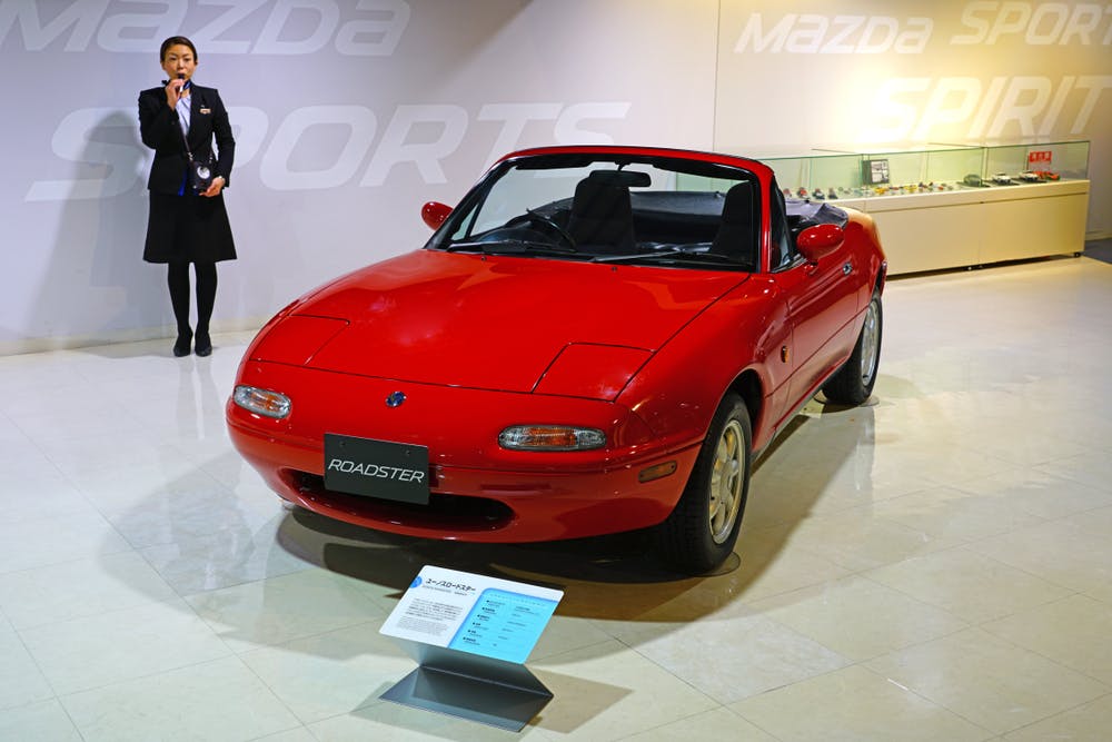 A red Mazda Roadster convertible is displayed indoors on a shiny floor, with an information plaque in front. A woman in a black suit stands nearby, speaking into a microphone.