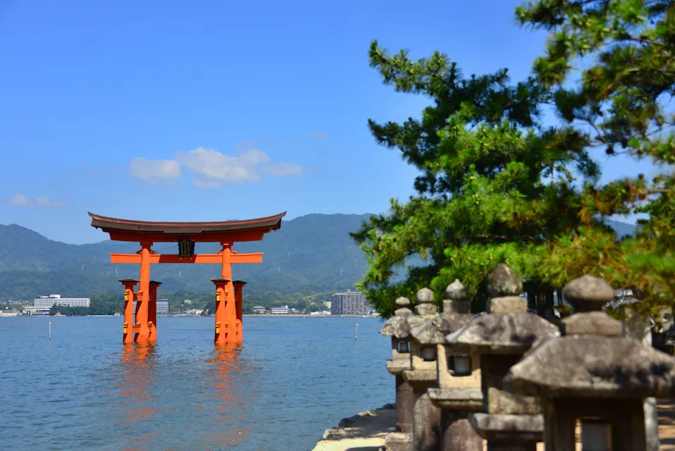 Miyajima Island 水辺に立つ鮮やかな赤い鳥居が、青い空と緑の山々の背景の中にあります。近くには、石灯籠と松の木が水辺に見えます。