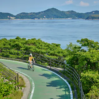 Shimanami Kaido A person wearing a helmet and yellow shirt rides a bicycle on a winding green path surrounded by lush greenery, with calm blue water and distant mountains in the background.