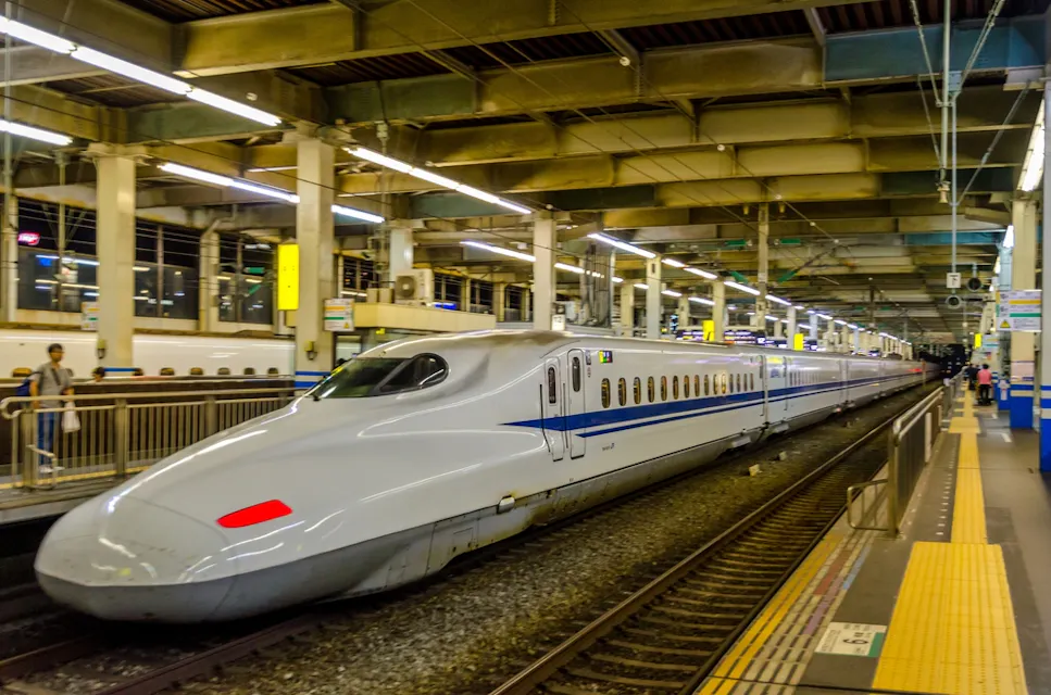 A sleek, modern bullet train is parked at a brightly lit station platform. The train is white with blue accents and features an aerodynamic design. The station is relatively empty, with a handful of people visible in the background.
