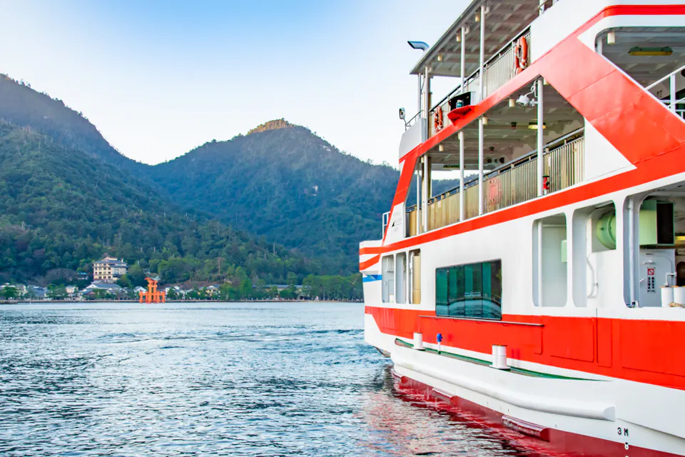A white and red ferry boat is sailing on a calm body of water with a mountainous landscape in the background. A prominent orange torii gate is visible on the water near the shore, adding a cultural landmark to the scenic view.