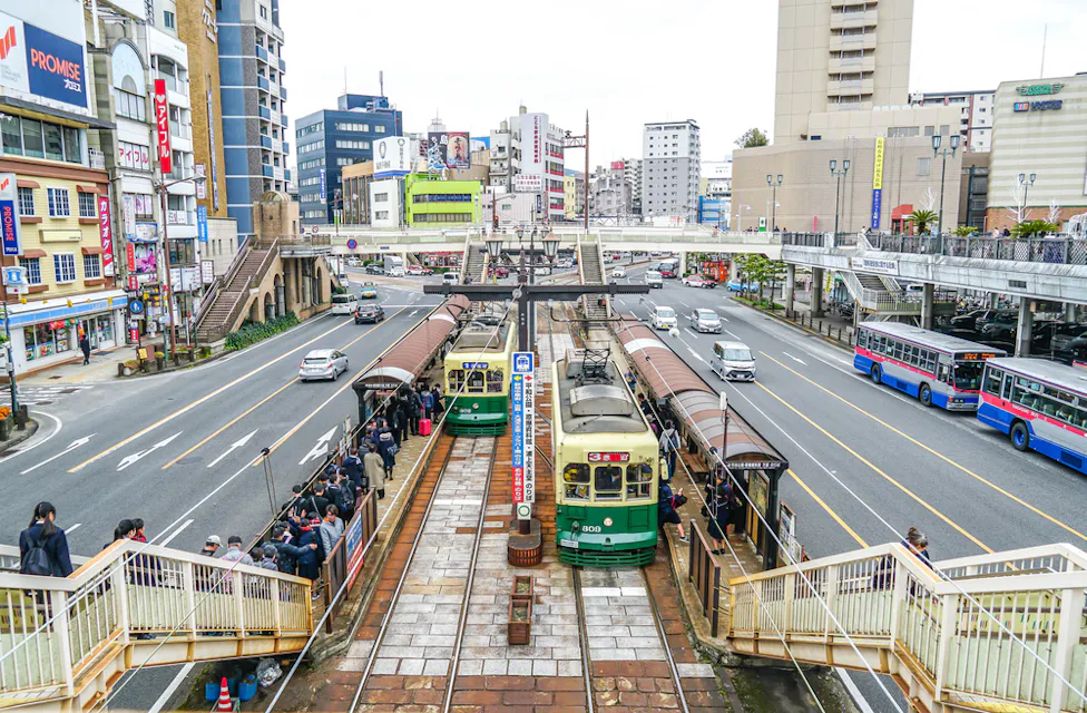 A bustling city scene shows two tram cars at a station with people boarding and waiting. Surrounding the station are multi-story buildings, shops, and a few buses on the road. Elevated walkways connect different parts of the area, and vehicles are seen on the streets.