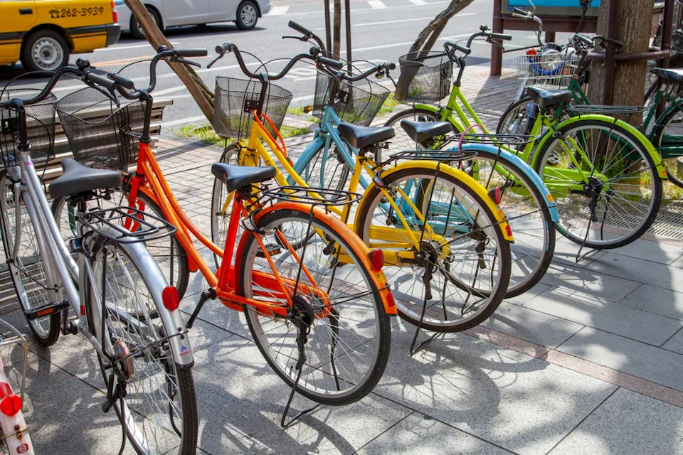 A row of colorful bicycles, including orange, yellow, blue, and green, parked on a paved sidewalk. Baskets are attached to the front of the bicycles. Trees and a passing car are visible in the background.