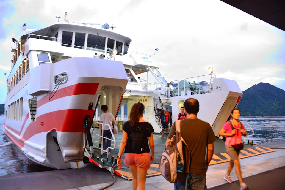 People boarding a large white and red ferry docked at a terminal. The vessel has ramps lowered for passenger access. A mountainous landscape and water are visible in the background. The sky is partly cloudy, suggesting it might be late afternoon or early evening.
