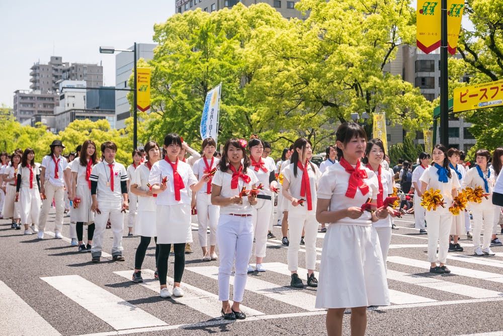 A group of people dressed in white with red scarves march in formation on a sunny city street, some holding pom-poms and musical instruments, with green trees and buildings in the background.