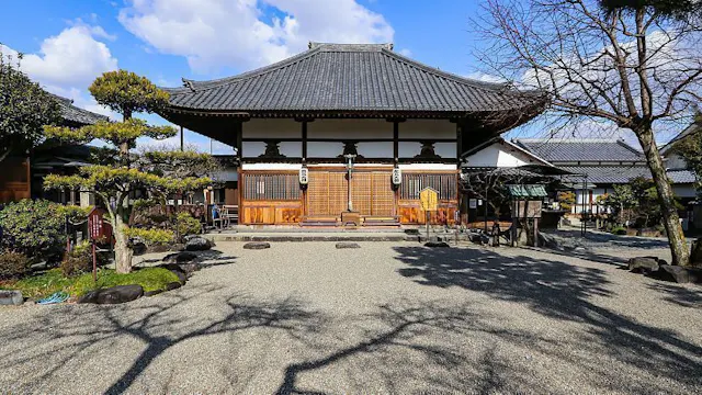 Traditional Japanese temple with a tiled roof, wooden sliding doors, and paper lanterns, surrounded by manicured trees and gravel courtyard under a sunny, partly cloudy sky.