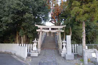 A traditional Japanese torii gate stands at the base of stone steps leading up to a shrine, surrounded by trees and stone lanterns, with informational signs on the right.