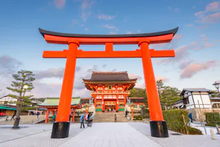 Large red torii gate stands at the entrance of Fushimi Inari Taisha shrine in Kyoto, Japan, with people walking and the main shrine building visible under a clear blue sky with scattered clouds.