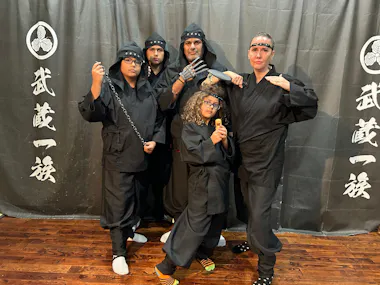A group of five people dressed as ninjas in black uniforms stands in front of a dark backdrop with Japanese characters. Two of them hold weapons, while others strike various poses. The backdrop has Japanese symbols and text.