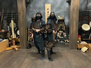 Two people dressed as ninjas are standing back-to-back in a room decorated with various traditional Japanese weapons and armor. They wear black and brown attire with face coverings, and the person on the left holds a sword. The room has a wooden floor and dark walls.