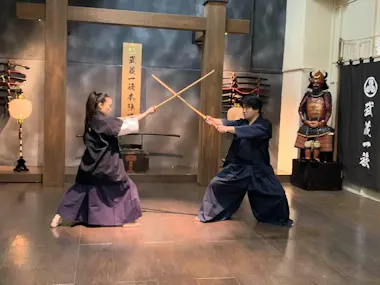 Two individuals are engaged in a kendo practice in a traditional dojo. Both wear black martial arts uniforms and face each other with wooden swords (bokken) crossed. The dojo features samurai armor, a calligraphy banner, and decorative weapons on the wall.