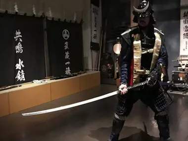 A person dressed in traditional samurai armor stands indoors, holding a long katana sword in a defensive stance. The backdrop features black banners with white Japanese kanji and various traditional items. The setting appears to be a museum or historic exhibit.
