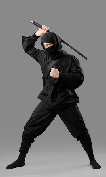 Ninja A person dressed in a black ninja outfit, including a mask that covers most of their face, stands in a dynamic pose with one foot forward. They are holding a katana sword raised above their head, ready to strike. The background is a plain gray.