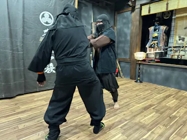 Two people dressed in black martial arts uniforms and face coverings are engaged in a sparring session in a dojo with wooden floors. One person has their arm raised while the other person steps back. Traditional martial arts weapons and gear are visible in the background.