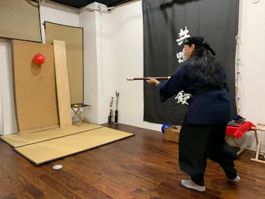 A person wearing traditional martial arts attire performs a blowgun demonstration in a room with wooden floors and Japanese decor. They aim at a red balloon fixed to a vertical wooden board. A black banner with white Japanese characters hangs on the wall behind them.