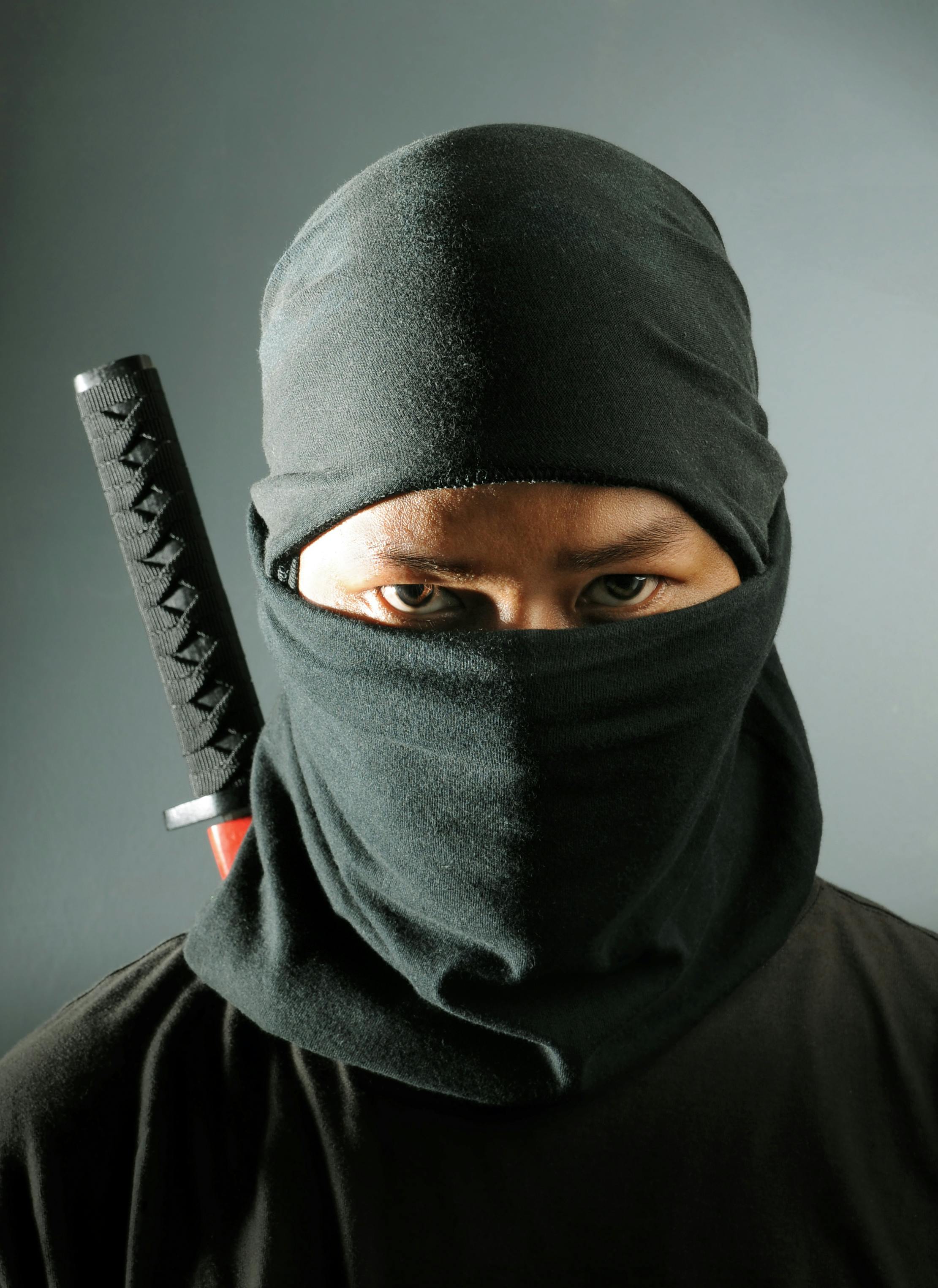 A person dressed as a ninja, wearing a black hood and mask that cover most of their face, with only their eyes visible. A sword is seen strapped on their back. The background is plain and gray.