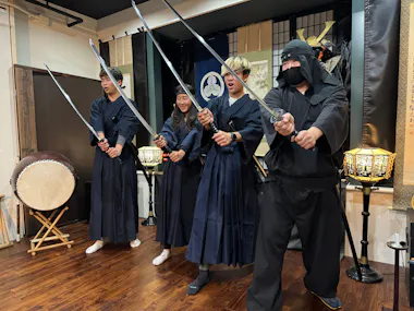Four people dressed in traditional samurai and ninja attire stand in a row, each holding a katana sword. They appear to be posing or taking part in a performance. There is a large drum and ornate lamps in the background, creating a historical Japanese setting.