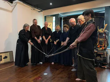A group of seven people stands in a semicircle, each holding a samurai sword extended towards the center, smiling and dressed in traditional martial arts attire. Behind them is a display with a Samurai armor set. The room appears to be a dojo with wooden flooring.