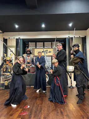 Five individuals dressed in traditional Japanese samurai attire pose with swords inside a room decorated with samurai armor and Japanese artifacts. They stand on a wooden floor, striking various poses with serious expressions.