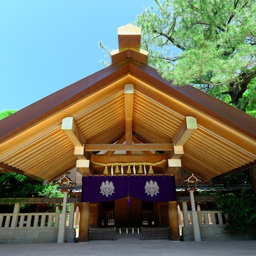 Atsuta-Jingu Shrine A traditional wooden Japanese Shinto shrine with a sharp-peaked roof stands amidst lush green trees. The entrance is adorned with purple banners featuring a white emblem, and a blue sky is visible in the background.