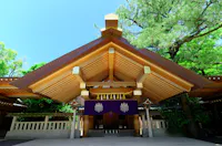 A traditional wooden Japanese Shinto shrine with a sharp-peaked roof stands amidst lush green trees. The entrance is adorned with purple banners featuring a white emblem, and a blue sky is visible in the background.