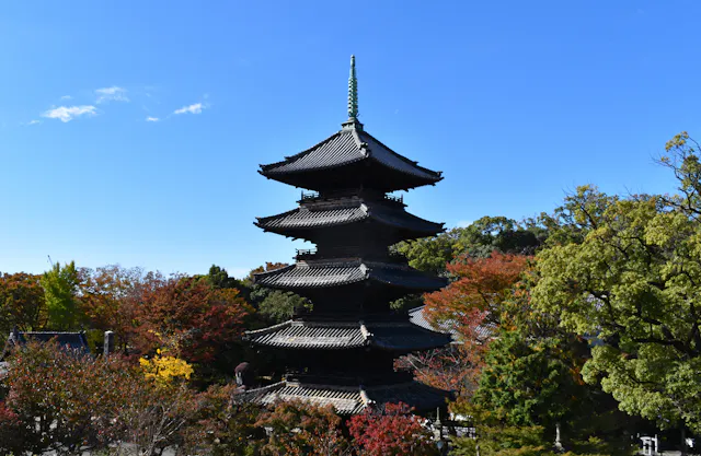 A traditional five-story Japanese pagoda stands among colorful autumn trees under a clear blue sky.