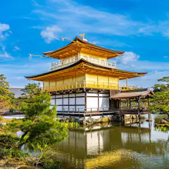 Kinkakuji A golden, multi-story Japanese pavilion sits beside a peaceful pond surrounded by lush trees and blue sky, reflecting beautifully on the water.