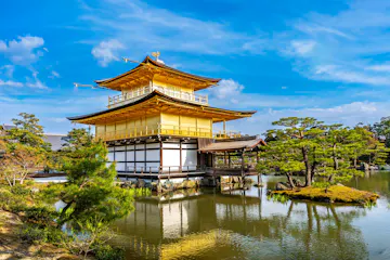 Kinkakuji A golden, multi-story Japanese pavilion sits beside a peaceful pond surrounded by lush trees and blue sky, reflecting beautifully on the water.