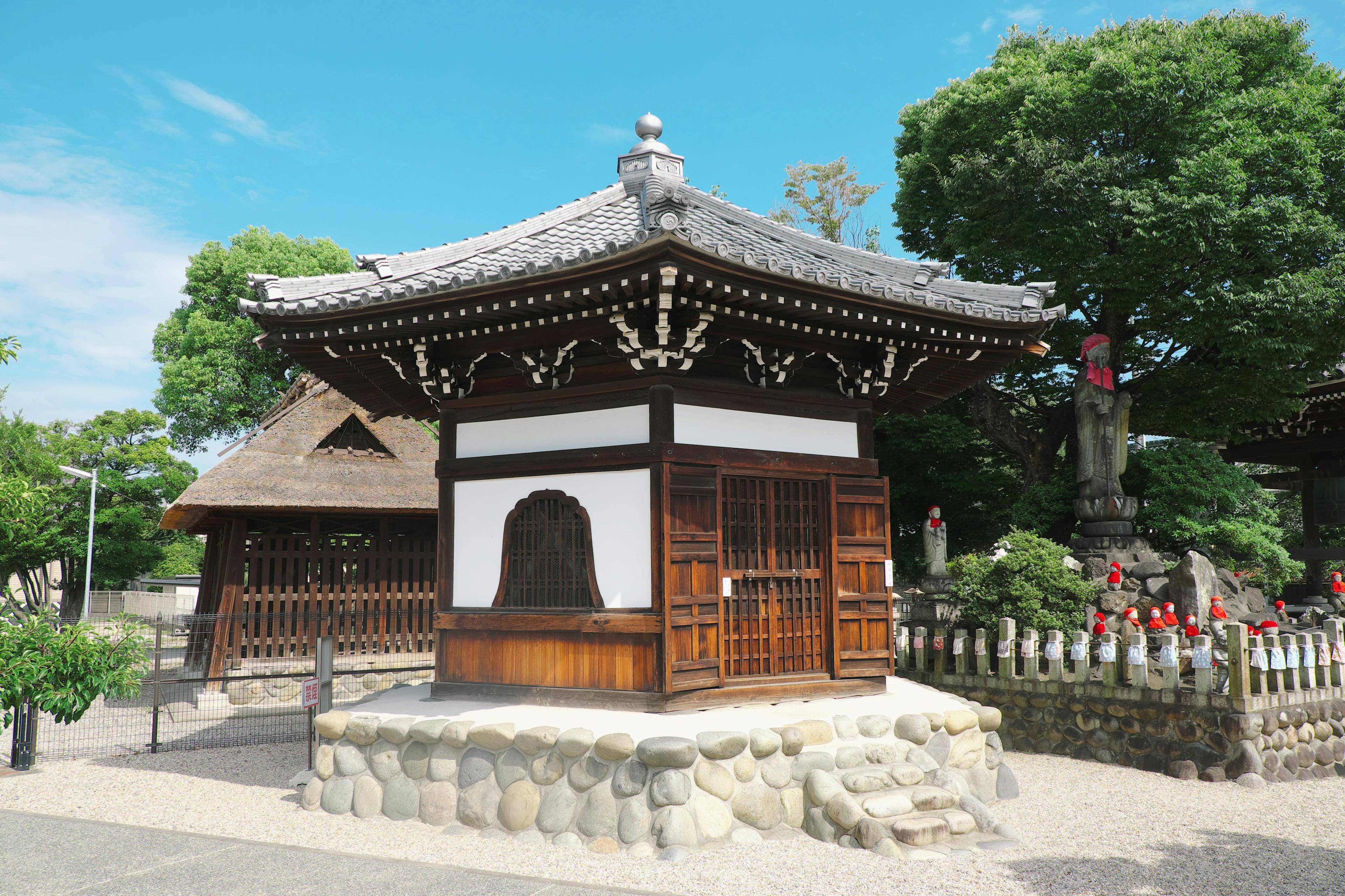 A small traditional Japanese wooden building with a tiled roof sits on a stone foundation, surrounded by trees and other structures under a clear blue sky.