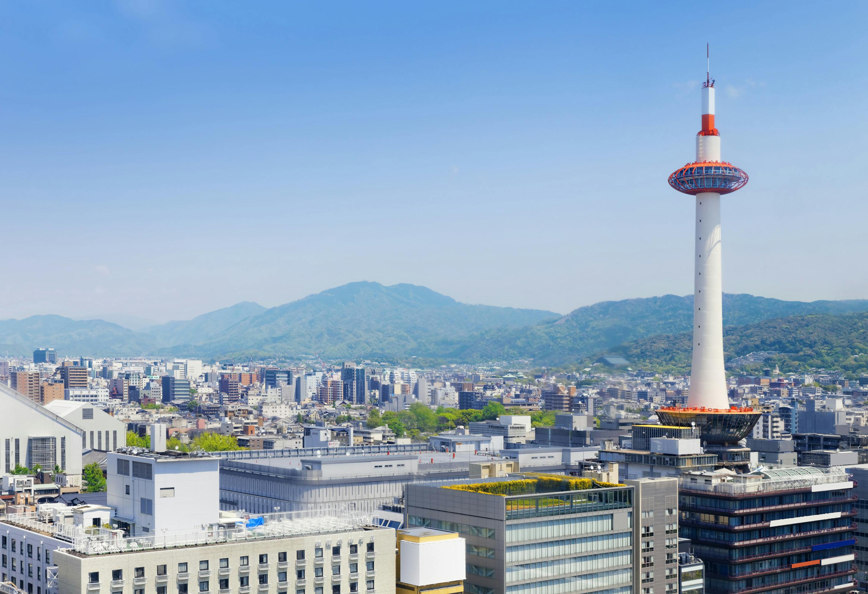 Kyoto cityscape with Kyoto Tower in the foreground, surrounded by modern buildings, and lush green mountains in the background under a clear blue sky.