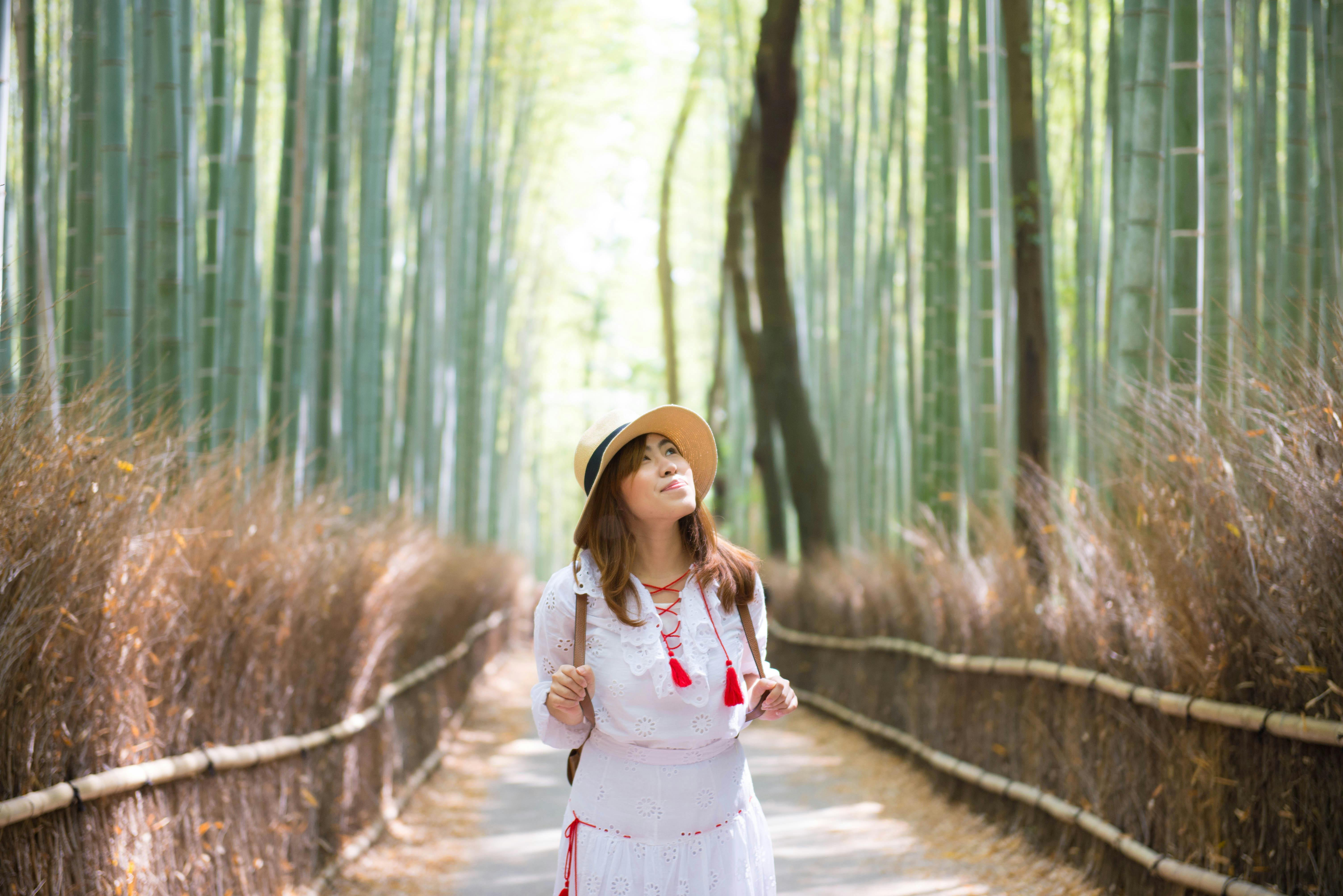 A woman wearing a white dress and straw hat walks along a path through a tall bamboo forest, looking up and smiling, with sunlight filtering through the trees.